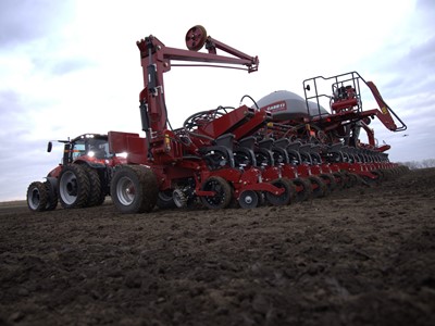 A large red agricultural tractor attached to a multi-row planter machine working on a freshly tilled field under a cloudy sky, preparing the soil for planting crops.