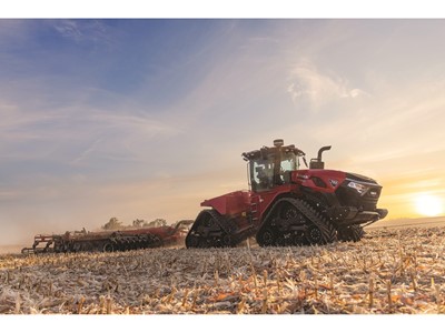 A large red Case IH tractor with rubber tracks is working in a harvested field during sunset, pulling farming equipment behind it under a partly cloudy sky.