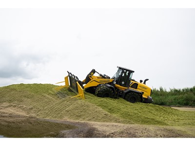 A yellow New Holland W170D wheel loader is pushing and leveling a large pile of green silage on a farm, with a cloudy sky in the background.