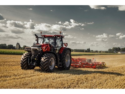 A red Case IH tractor with large tires is working in a golden wheat field under a partly cloudy sky, pulling a red agricultural implement for tilling the soil, with green crops and trees visible in the background.