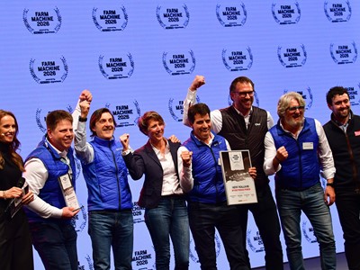 A group of eight people standing on stage celebrating an award at the Farm Machine 2026 event. Most of them are wearing blue vests and smiling, with some raising their fists in excitement. One person is holding a framed certificate that reads "New Holland Methane Power." The background features a repeated "Farm Machine 2026" logo.