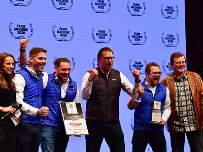A group of six people standing on stage celebrating an award. Four men in blue and black vests are holding plaques and certificates, smiling and raising their fists in excitement. A woman on the left holds a microphone and a man on the right wears a brown leather jacket and patterned shirt. The background displays multiple "Farm Machine 2026" logos.