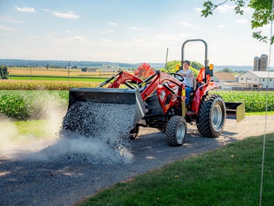 A person operating a red tractor with a front loader, spreading gravel on a rural dirt road. The background features green fields, a farmhouse, silos, and a clear blue sky with scattered clouds.