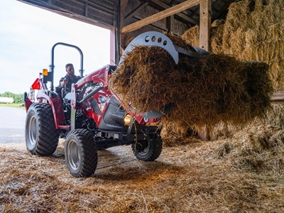 A person operating a red tractor inside a barn, using a front loader attachment to lift a large bale of hay. The barn is filled with stacked hay bales and straw scattered on the ground.