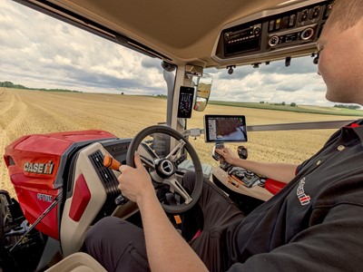 Interior view of a modern Case IH Optum tractor cabin showing a person operating the steering wheel and control panel, with a large golden wheat field visible through the windows under a cloudy sky.
