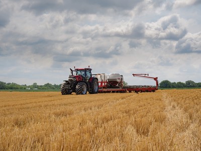 A red tractor with large tires is working in a golden wheat field under a cloudy sky, pulling farming equipment used for planting or fertilizing. Trees and greenery are visible in the distant background.