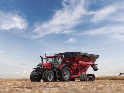 A red tractor attached to a large red grain cart with black tracks is parked in a harvested wheat field under a partly cloudy blue sky.