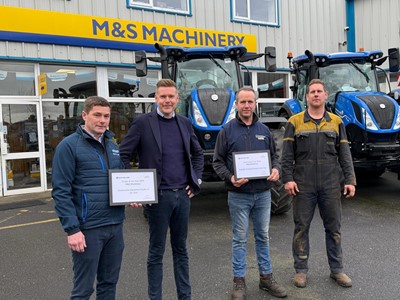Four men standing in front of two blue tractors outside a building with a yellow and blue "M&S Machinery" sign. Two of the men are holding framed certificates, and all are dressed in casual or work attire. The scene appears to be an award presentation or recognition event.