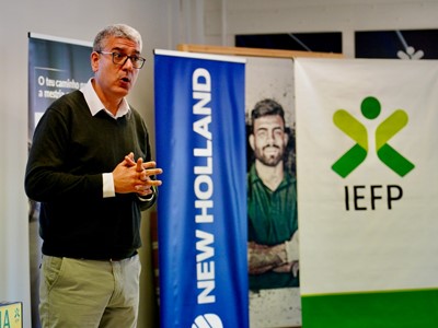 A man with gray hair and glasses, wearing a black sweater over a white collared shirt and beige pants, is speaking or presenting indoors. Behind him are three vertical banners, one blue with the text "NEW HOLLAND," another with a photo of a man with crossed arms, and a white banner with a green and yellow logo and the text "IEFP.