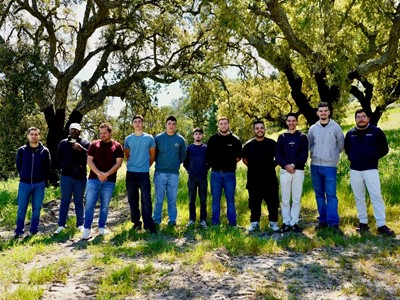 A group of ten young men standing outdoors in a sunny, grassy area with large trees in the background, all facing the camera and posing for a group photo.