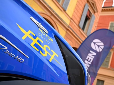 Close-up of a blue vehicle with yellow text "Motor Valley Fest" and model designation "T7.340 HD" on its side, set against a background of colorful buildings and a blurred flag with the word "New" visible.