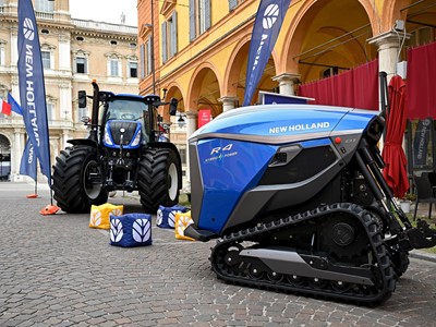 A blue New Holland R4 Hybrid Power autonomous agricultural vehicle with tracks is displayed on a cobblestone street next to a traditional blue New Holland tractor with large tires. The scene is set in an urban area with historic buildings and arches in the background, accompanied by New Holland branded flags and colorful cube seats on the ground.