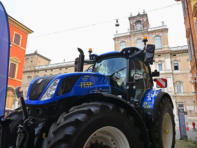 Blue New Holland tractor with "Motor Valley Fest" branding displayed in an urban setting with historic buildings in the background, under a clear sky.