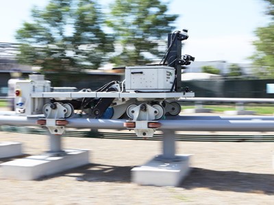 A mobile robot equipped with cameras and sensors is moving along a metal rail outdoors, with blurred trees and buildings in the background indicating motion.
