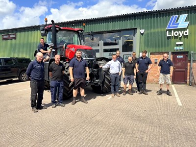 A group of nine men standing and posing in front of a large red tractor outside a green industrial building with the sign "Lloyd" on it. One man is sitting on the tractor, while the others stand on the ground, smiling and dressed in casual work attire. The scene is set on a sunny day with a blue sky and some clouds.
