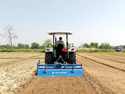 A farmer driving a white New Holland tractor with a blue tiller attachment, plowing a large, dry agricultural field under a clear sky, with green trees and other tractors visible in the background.