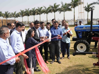 A group of people gathered outdoors on a grassy area with palm trees in the background, participating in a ribbon-cutting ceremony. A woman and a man in the center are cutting a red ribbon with scissors, while others stand around them watching. A blue New Holland tractor is visible on the right side of the image. The event appears to be a formal or celebratory occasion.