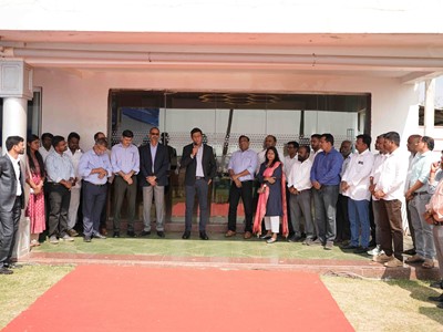 A group of professionally dressed men and women standing in a semi-circle outside a building with a red carpet laid out in front. One man in the center is speaking into a microphone, suggesting a formal event or gathering. The background shows glass doors and a white exterior wall.