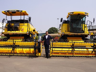 A man in a dark suit standing between two large yellow New Holland agricultural combine harvesters parked outdoors on a paved surface, with a building and greenery in the background under a clear sky.