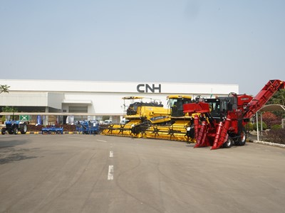Outdoor display of large agricultural machinery including yellow combine harvesters and a red corn harvester parked in front of a white industrial building with the CNH logo, under a clear blue sky.