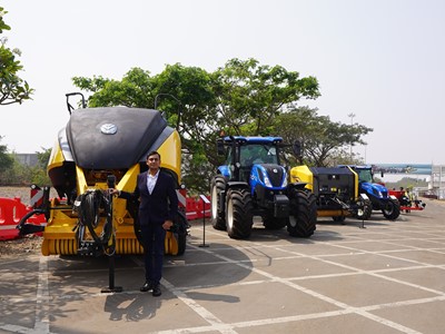 A man in a dark suit stands next to large yellow and black agricultural machinery and blue tractors parked in a row on a paved outdoor area with trees and red barriers in the background under a clear sky.