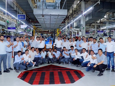 A large group of factory workers and engineers posing together inside a manufacturing facility, all giving thumbs-up gestures. They are standing and squatting around a blue tractor assembly line area, with industrial equipment and machinery visible in the background. The environment is well-lit with overhead lights and organized workstations.