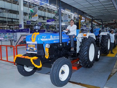 A man sitting on a blue New Holland TX 3600-2 tractor decorated with orange marigold garlands inside a factory assembly line. The tractor has a sign indicating "800000" units produced. Several similar tractors are lined up behind it, and factory workers are visible in the background. The setting is a well-lit industrial environment with various signs and equipment.