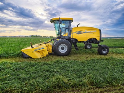 A yellow New Holland agricultural harvester machine is parked on a green field under a partly cloudy sky during daylight, with freshly cut crops visible on the ground.