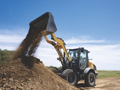 A yellow New Holland wheel loader is dumping a load of dirt onto a large pile at a construction site under a clear blue sky.