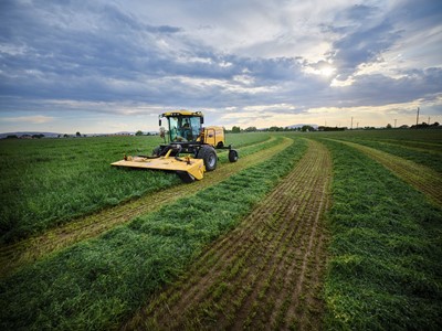 A yellow agricultural tractor mowing a large green field under a cloudy sky with the sun partially visible, creating neat rows of cut grass on the farmland.