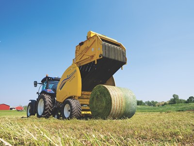 A blue tractor attached to a yellow New Holland round baler machine is operating in a green field under a clear blue sky, producing a large round bale of hay. In the background, there are some trees and a red barn.