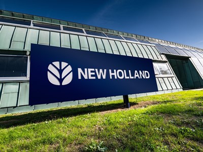 Insegna blu con il logo e la scritta "NEW HOLLAND" davanti a un edificio moderno con pareti in metallo verde e finestre, su un prato verde sotto un cielo sereno.