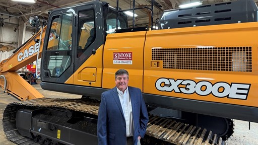 A man in a suit stands in front of a large orange Case CX300E excavator inside a warehouse. The excavator features a prominent logo and is positioned on a track, showcasing its size and design.