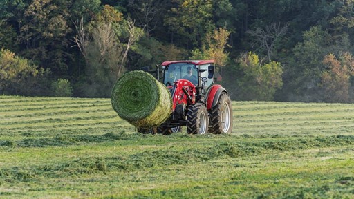 A red tractor pulling a large round hay bale across a lush green field, surrounded by trees and a clear sky. The scene captures the essence of agricultural work and rural life.