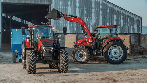 Two red tractors are parked in a farmyard. The tractor on the left has a front loader attachment and a driver inside, while the tractor on the right is equipped with a rear implement. In the background, a large barn with a metal roof is visible, and the ground is dusty, indicating an agricultural setting.