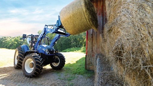 A blue tractor with a front loader is lifting a large round hay bale near a barn. The tractor is positioned on a gravel surface with a grassy field in the background, under a clear blue sky.