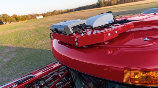 A close-up view of the top of a red agricultural machine, featuring mounted equipment including sensors and communication devices. The background shows a green field under a clear sky, indicating a rural farming environment.