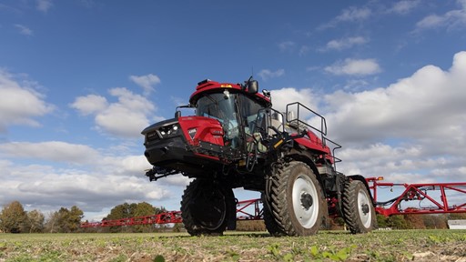 A red agricultural sprayer vehicle parked on a field under a partly cloudy sky. The sprayer features large wheels, a high cabin, and is equipped for crop spraying, showcasing its modern design and functionality.