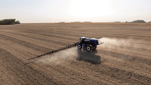 A blue tractor equipped with a sprayer is working in a vast, empty agricultural field. Dust is being kicked up as the tractor moves across the dry, brown soil under a clear sky. The landscape is flat and stretches into the distance, showcasing the agricultural setting.