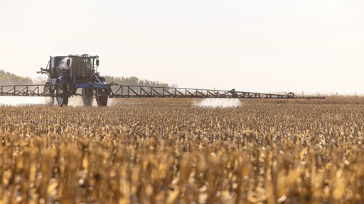 A large agricultural sprayer tractor applying chemicals to a field of dry, golden crops under a clear sky. The tractor is positioned in the distance, with a long spray arm extending outward, creating a mist as it moves through the harvested field.