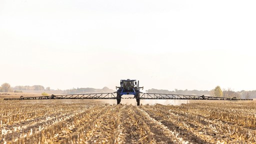 A large agricultural sprayer machine operates in a field, applying chemicals to the soil. The landscape features rows of harvested crops, with a clear sky in the background, indicating a sunny day.