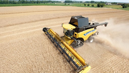 A yellow combine harvester working in a vast field of golden crops under a clear blue sky. Dust is being kicked up as the machine moves through the field, showcasing the agricultural landscape with green trees in the background.