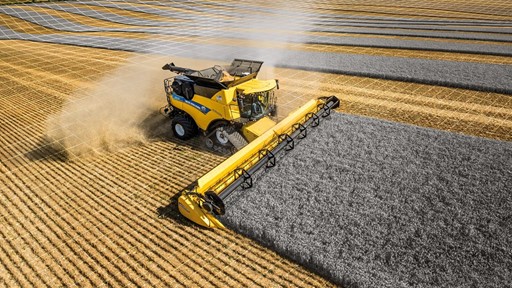 A yellow combine harvester is cutting through a field of golden crops, creating neat rows of harvested grain. The sky is clear with a few clouds, and the landscape features alternating stripes of harvested and unharvested fields, showcasing agricultural activity.