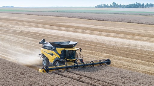 A yellow combine harvester is working in a vast field of golden wheat. The machine is cutting through the crops, with dust rising behind it. The landscape features a clear blue sky and distant trees, showcasing a rural agricultural setting.