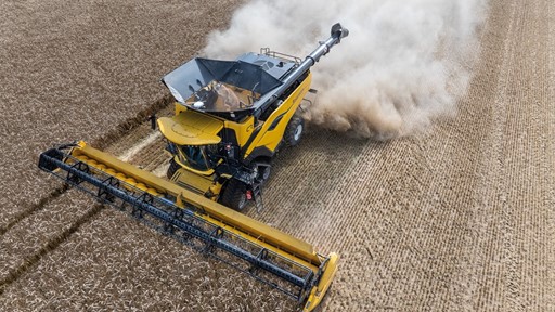 Aerial view of a yellow combine harvester working in a field, with dust and debris being kicked up as it cuts through rows of crops. The machine is equipped with a wide header, efficiently harvesting the agricultural produce. The surrounding field shows neatly arranged crop rows, highlighting the agricultural landscape.