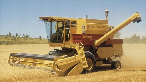 A New Holland TF70 combine harvester working in a golden wheat field, with dust rising as it cuts through the crops under a clear blue sky.