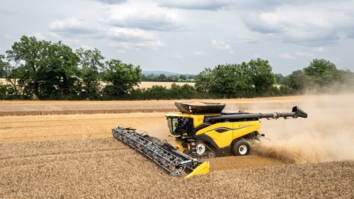 A yellow and black combine harvester is working in a field, harvesting golden wheat. Dust is being kicked up as the machine moves through the crop, with green trees and a cloudy sky in the background. The scene captures the essence of agricultural activity during the harvest season.