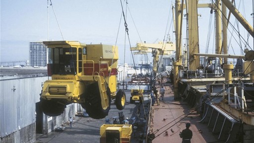 A yellow agricultural harvester being lifted by a crane at a port, with cargo ships and workers in the background. The scene depicts industrial activity, showcasing the transportation of heavy machinery.