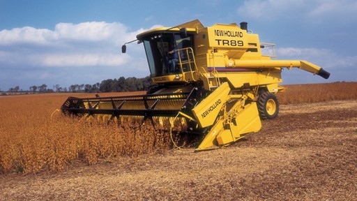A yellow New Holland TR89 combine harvester working in a soybean field under a blue sky with scattered clouds. The machine is cutting through tall crops, showcasing agricultural machinery in action.