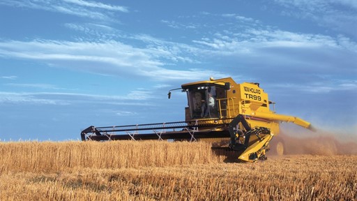 A yellow combine harvester working in a golden wheat field under a blue sky with scattered clouds. The machine is cutting through the tall wheat stalks, creating a cloud of dust.